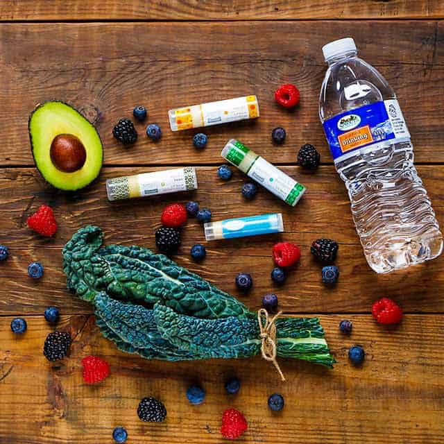 Greens, avocado, chapstick, bottled water and berries on wood table