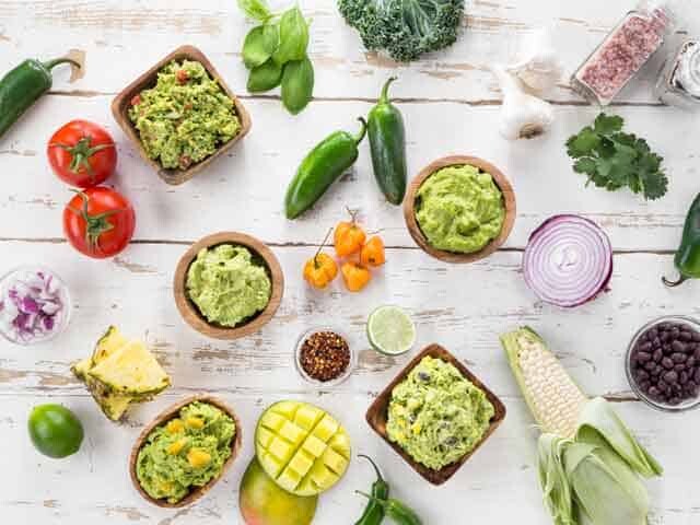 Bowls of Guacamole dips shown with all the fresh ingredients on white wood